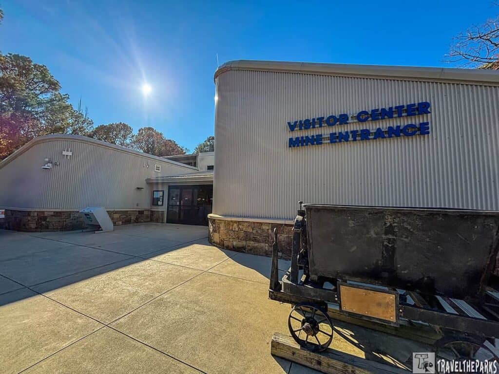 Visitor center with a mine entrance under a sunny blue sky, featuring a vintage mining cart in front.