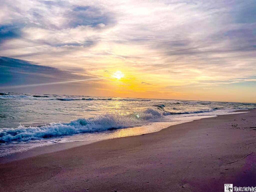 The horizon is adorned with a glowing sun partially hidden by a mix of clouds, casting a warm golden hue across the sky. The sky transitions from warm yellows and oranges near the sun to cooler blues and greys towards the edges. Gentle waves roll onto the sandy shore, reflecting hints of sunlight. The beach appears serene and unoccupied, with the sand stretching uninterrupted along the waterline.