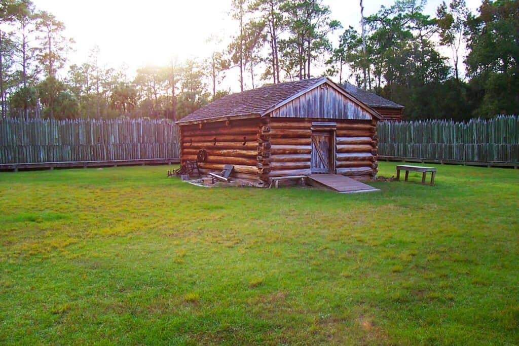 A log cabin with a wooden fence and trees in the background during sunset.