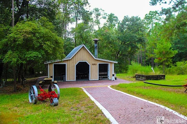 A small wooden building with a metal roof in a wooded area, with a brick path leading to it and a red vintage machine nearby.