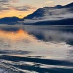 Sunset over a tranquil sea with mountain silhouettes and vibrant sky reflections.