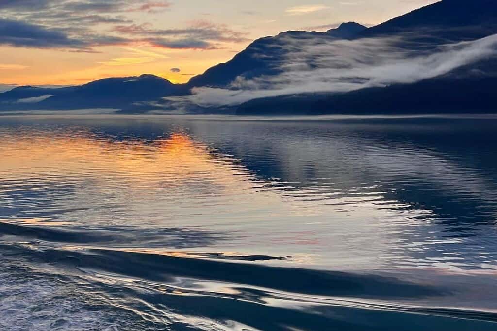 Sunset over a tranquil sea with mountain silhouettes and vibrant sky reflections.