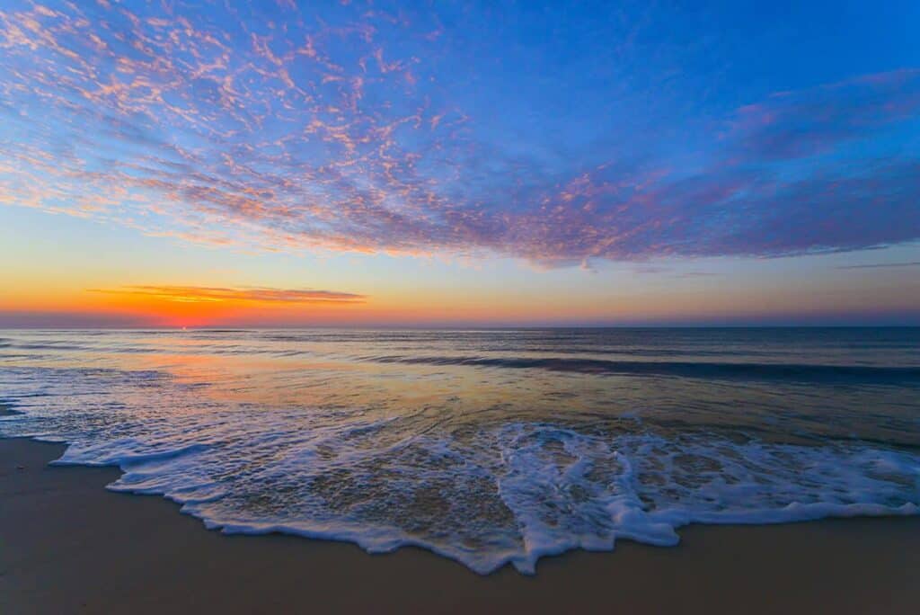 On the Emerald Coast the sky is filled with vibrant colors, transitioning from deep blue at the top to rich orange and yellow near the horizon where the sun is setting. Wispy clouds stretch across the sky, picking up hues of pink and purple. The gentle waves of the ocean extend toward the sandy shore, with white sea foam visible at the water's edge. The overall atmosphere is tranquil and picturesque.