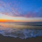 On the Emerald Coast the sky is filled with vibrant colors, transitioning from deep blue at the top to rich orange and yellow near the horizon where the sun is setting. Wispy clouds stretch across the sky, picking up hues of pink and purple. The gentle waves of the ocean extend toward the sandy shore, with white sea foam visible at the water's edge. The overall atmosphere is tranquil and picturesque.