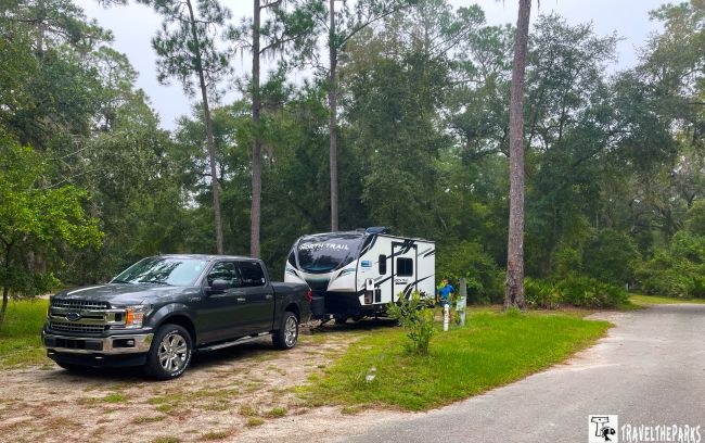 a campsite at Stephen Foster Folk Cultural State Park. In the foreground, there is a dark gray pickup truck parked next to a white travel trailer. The trailer has a modern design with black and dark blue accents. The vehicles are situated on a gravel area surrounded by lush greenery. Tall pine trees, with slender trunks and sparse foliage, dominate the background, creating a natural canopy. The ground is a mix of green grass and patches of bare earth. A paved road curves away from the campsite to the right. The setting appears quiet and serene, typical of a state park environment.