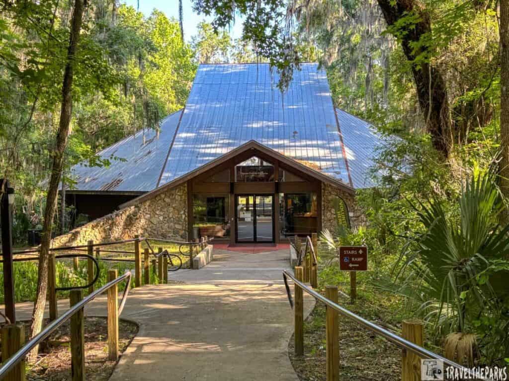 Entrance of Paynes Prairie Preserve State Park Visitor Center, with a metal roof, stone facade, and glass doors, surrounded by greenery.