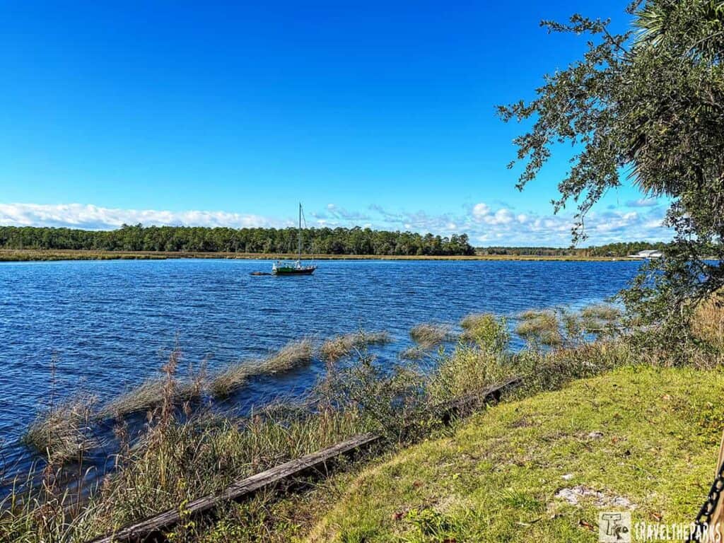 A view of the Wakulla River with a sailboat, grassy bank, and tree-lined horizon under a clear blue sky.