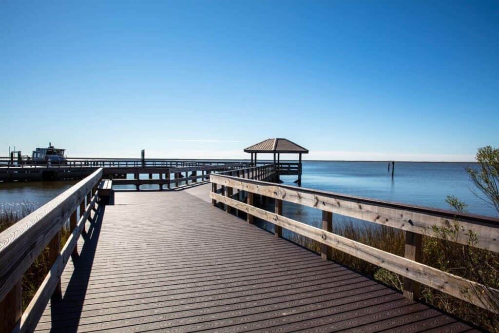 a scenic wooden boardwalk extending across a coastal bayou. It features a wide pathway made from evenly laid wooden planks that lead towards a simple, open-sided gazebo with a pyramidal, dark brown roof, located over the calm water. To the left, a boat is docked along a connected pier. The right side of the image shows the clear, blue water of the bayou, bordered by marsh grasses and scattered shrubs. The sky above is clear and bright blue, adding to the tranquil setting.