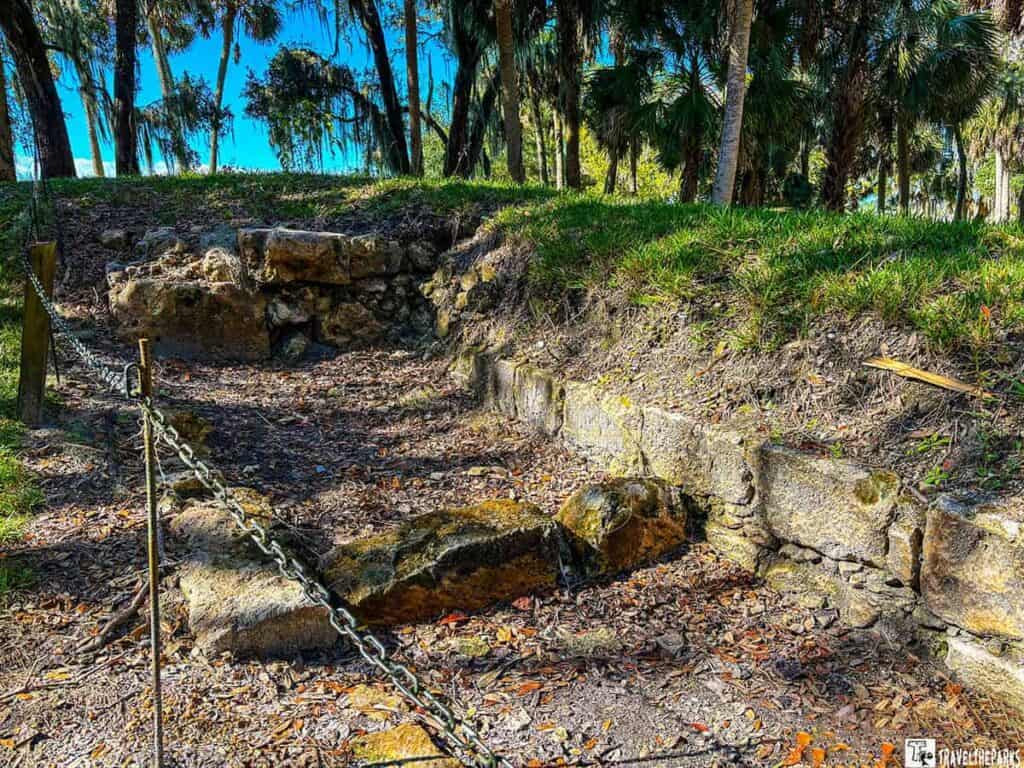 The stone remaining walls of rooms with leaf-covered ground at Fort San Marcos de Apalache, bordered by lush trees and a chain barrier.
