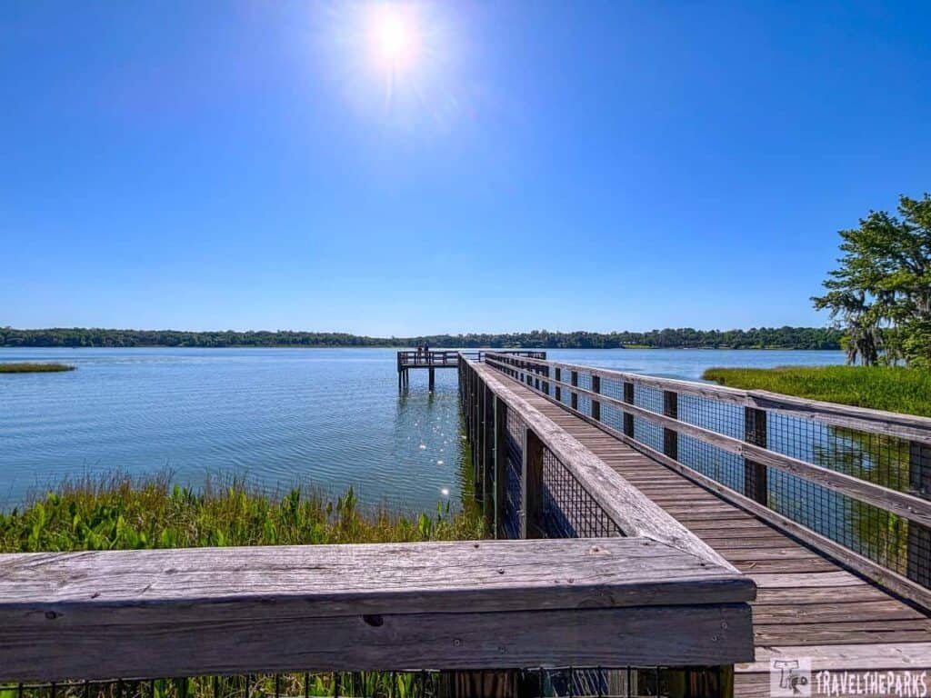 A wooden pier extends into a tranquil lake under a bright sun with trees and greenery around.