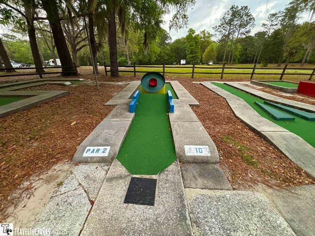 Mini-golf hole with a red tunnel obstacle and blue barriers, surrounded by trees and a wooden fence.