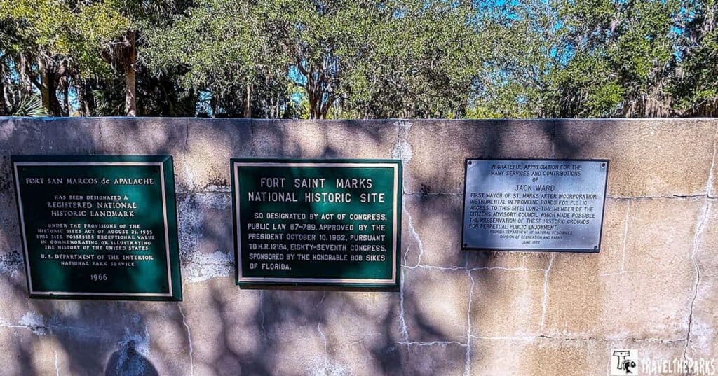 Three plaques on a concrete wall, commemorating Fort San Marcos de Apalache, Fort Saint Marks National Historic Site, and Jack Ward's contributions, with trees in the background.