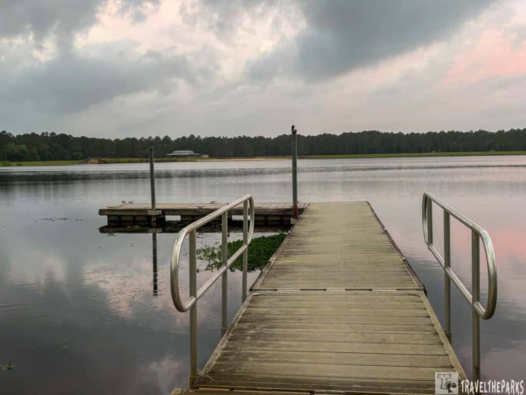 A wooden dock with metal railings extending over a calm body of water. The dock leads towards an adjacent small platform on the left. The water is reflective, capturing the overcast sky which is filled with gray clouds tinged with soft pink hues. Across the water, a line of dense trees stretches along the horizon. There is a structure visible in the distance on the opposite shore. The foreground includes some greenery floating in the water near the dock.