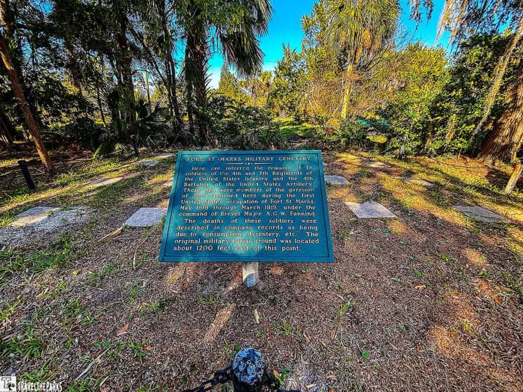 Green historical plaque at Fort St. Marks Military Cemetery surrounded by trees and grave markers.