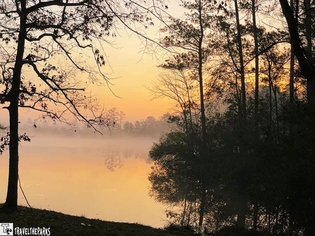 A serene lakeside scene at sunrise or sunset, with mist over the water and trees silhouetted against an orange sky.