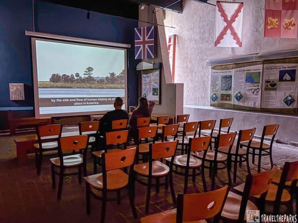Small museum auditorium with rows of wooden chairs facing a projection screen showing a landscape. Historical informational panels and flags are on the walls.