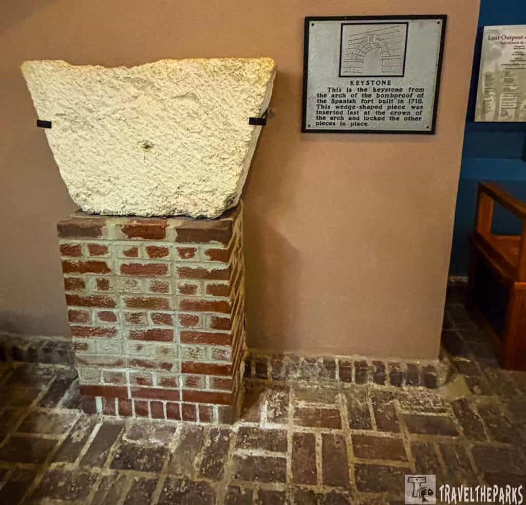 A keystone displayed on a brick pedestal with an informational plaque on the wall beside it.