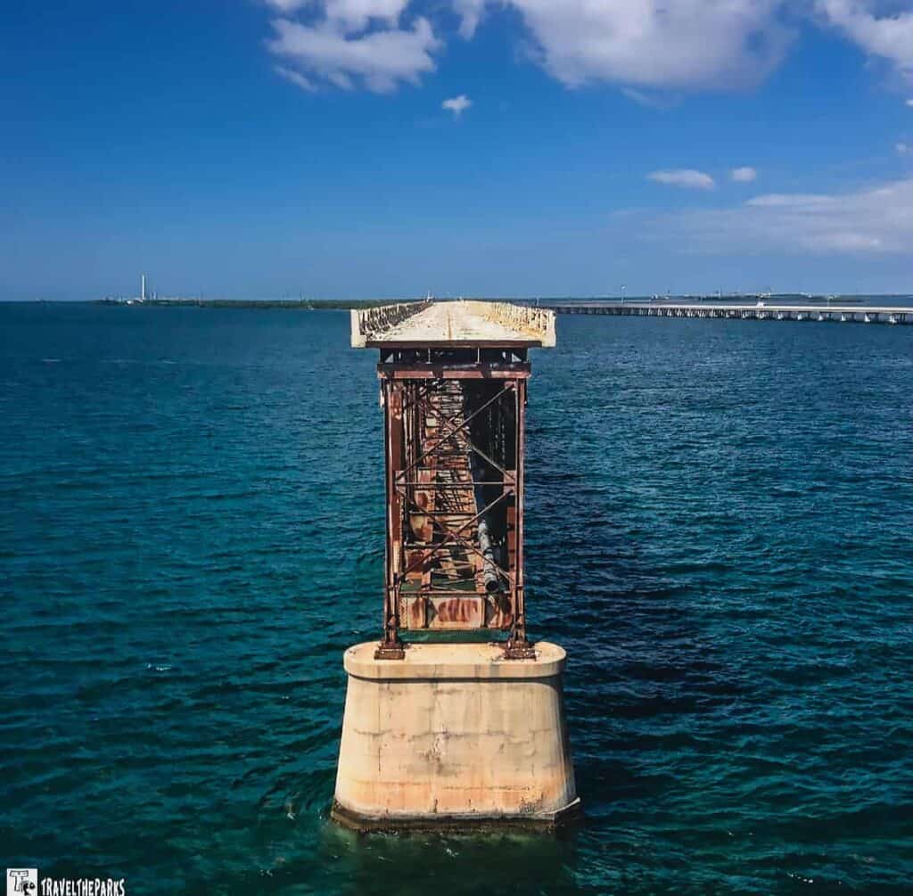 Rusted metal bridge support in the ocean with distant land and a bridge in the background.