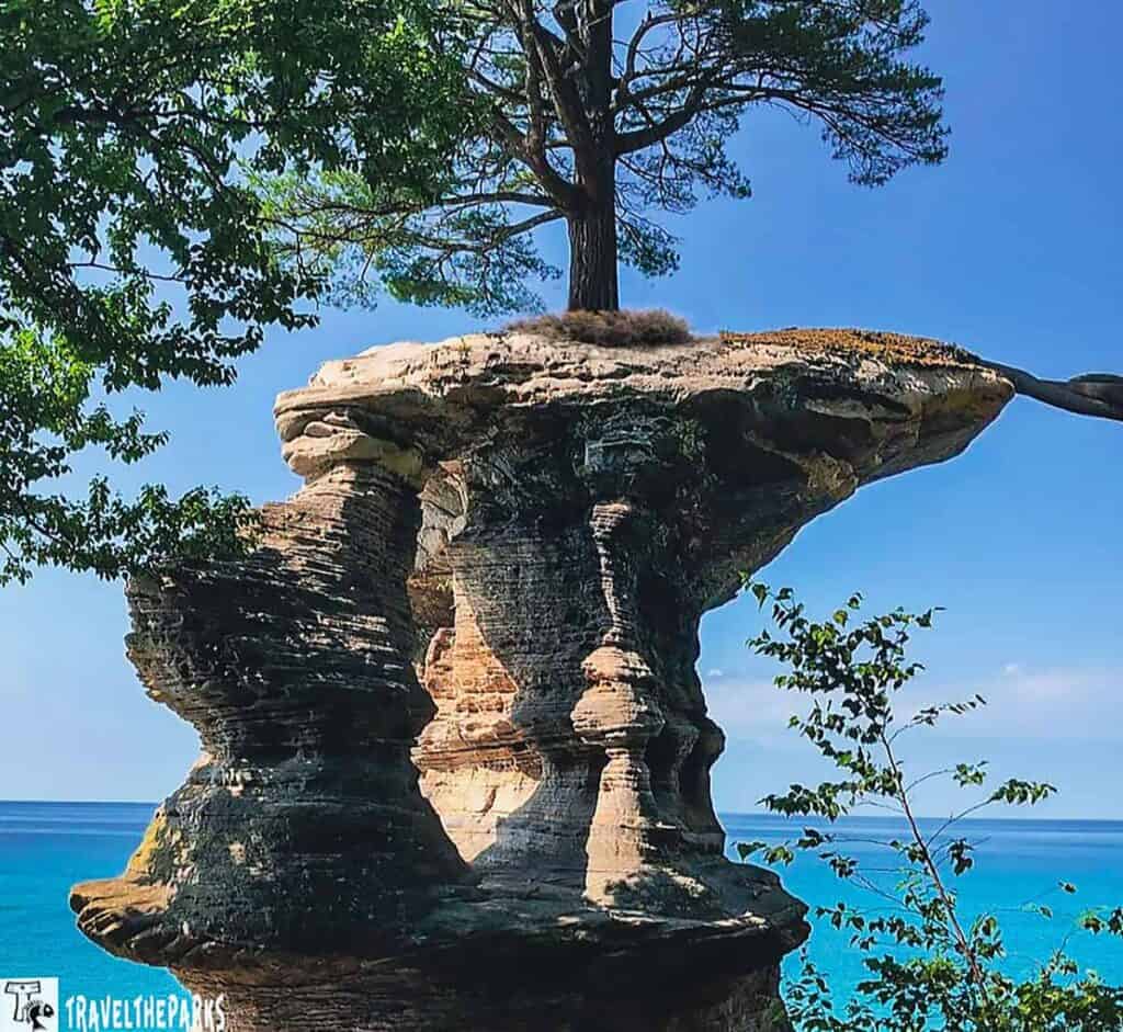 Chapel Rock with a pine tree on top at Pictured Rocks National Lakeshore, set against a blue sky and lake.