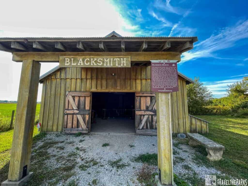 A rustic wooden blacksmith building with an open entrance under a blue sky, featuring a sign that reads "BLACKSMITH."