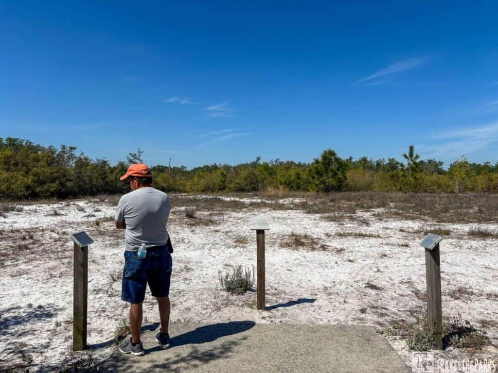 A man in a gray t-shirt and orange cap stands on a sandy path with vegetation in the background at the Fort Chokonikla site in Paynes Creek Historic State Park.