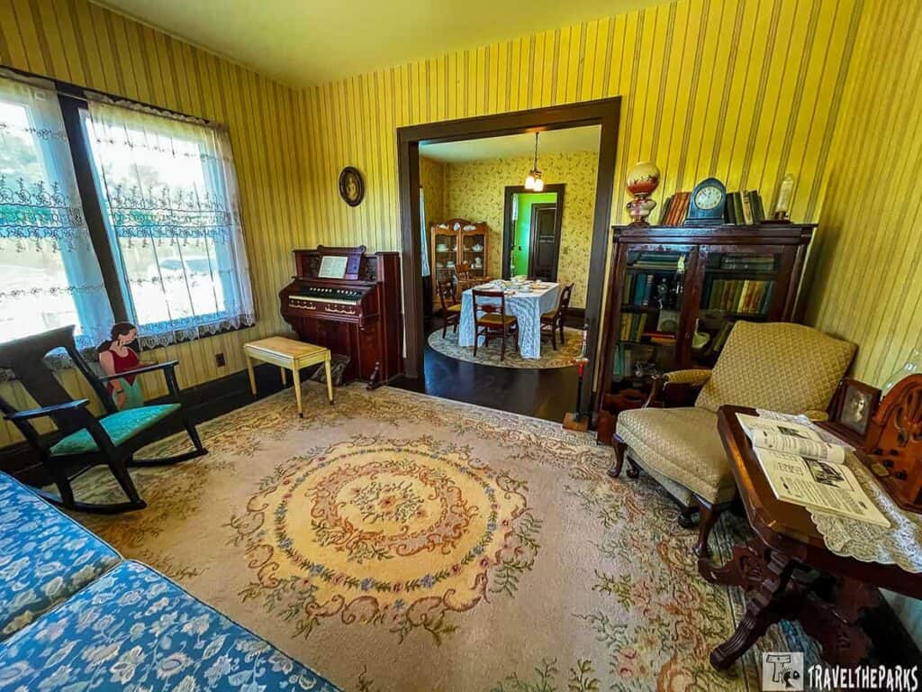 Vintage living room with striped wallpaper, floral carpet, organ, cabinet, and view into a dining room.