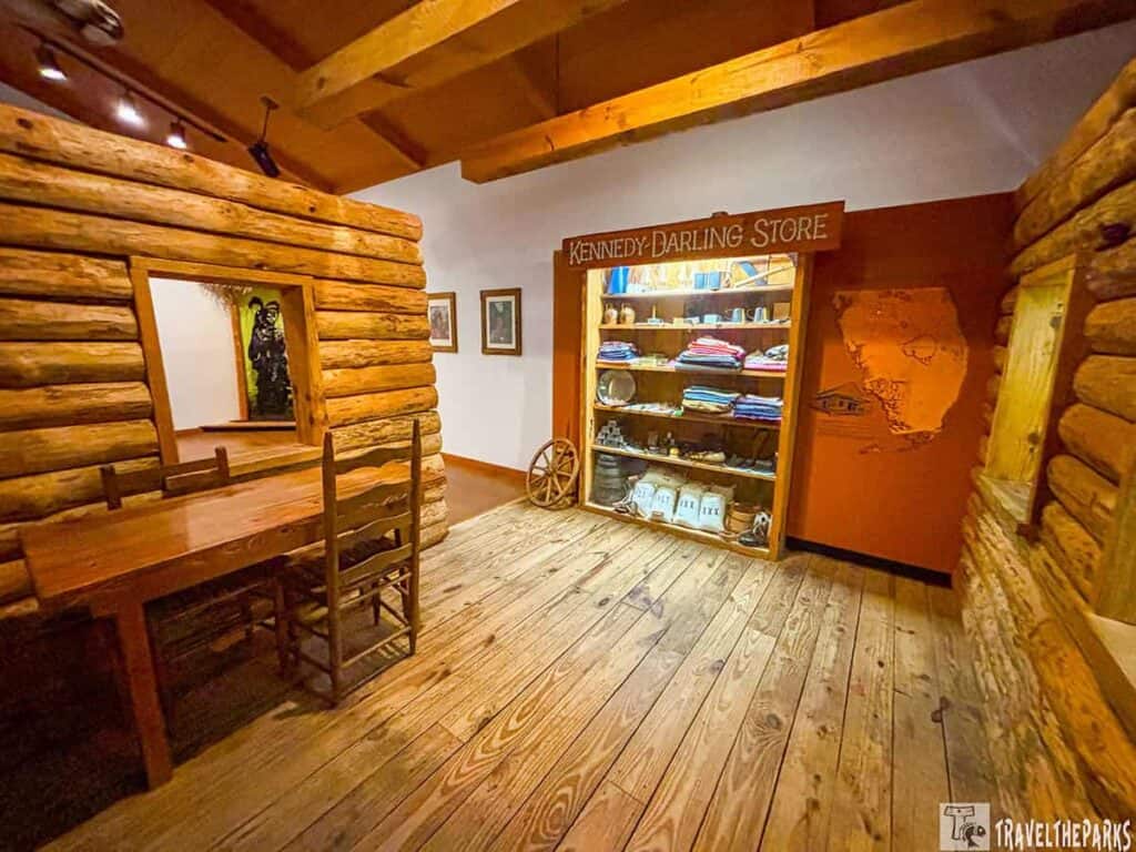 Interior of a cabin with logs, a wooden table, a display labeled "Kennedy-Darling Store," and an orange wall featuring a map of Florida.