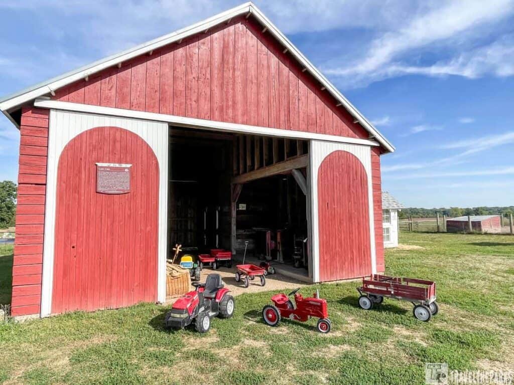  red wooden barn with large white-trimmed doors, partially open to reveal a dark interior. It stands on a grassy lawn under a blue sky scattered with thin clouds. In front of the barn, several small, red toy tractors and wagons are scattered on the grass. To the right, a more extensive field and another small structure are visible in the background.