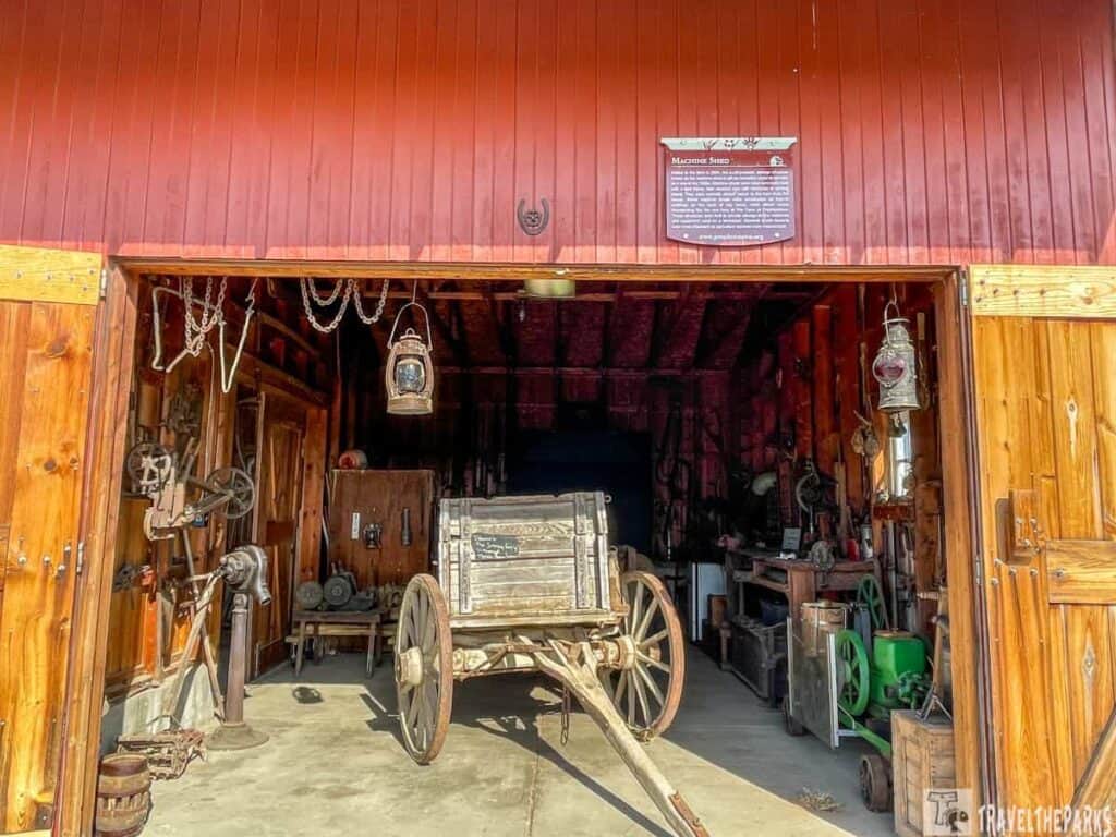 Open entrance of a red wooden machine shed with vintage machinery and a wooden wagon inside.