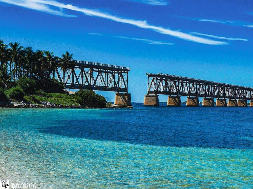 An abandoned Bahia Honda rail bridge over blue ocean waters with palm trees on the left and clear skies with wispy clouds.