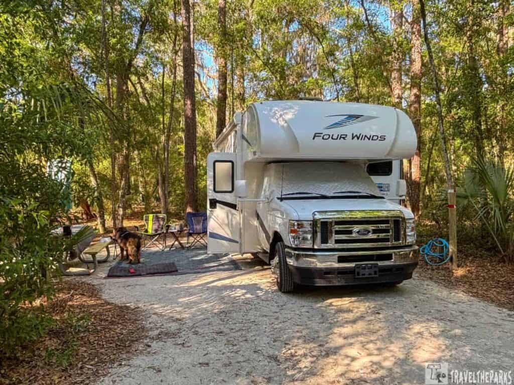 A "Four Winds" motorhome parked at a wooded campsite with outdoor chairs and a dog.