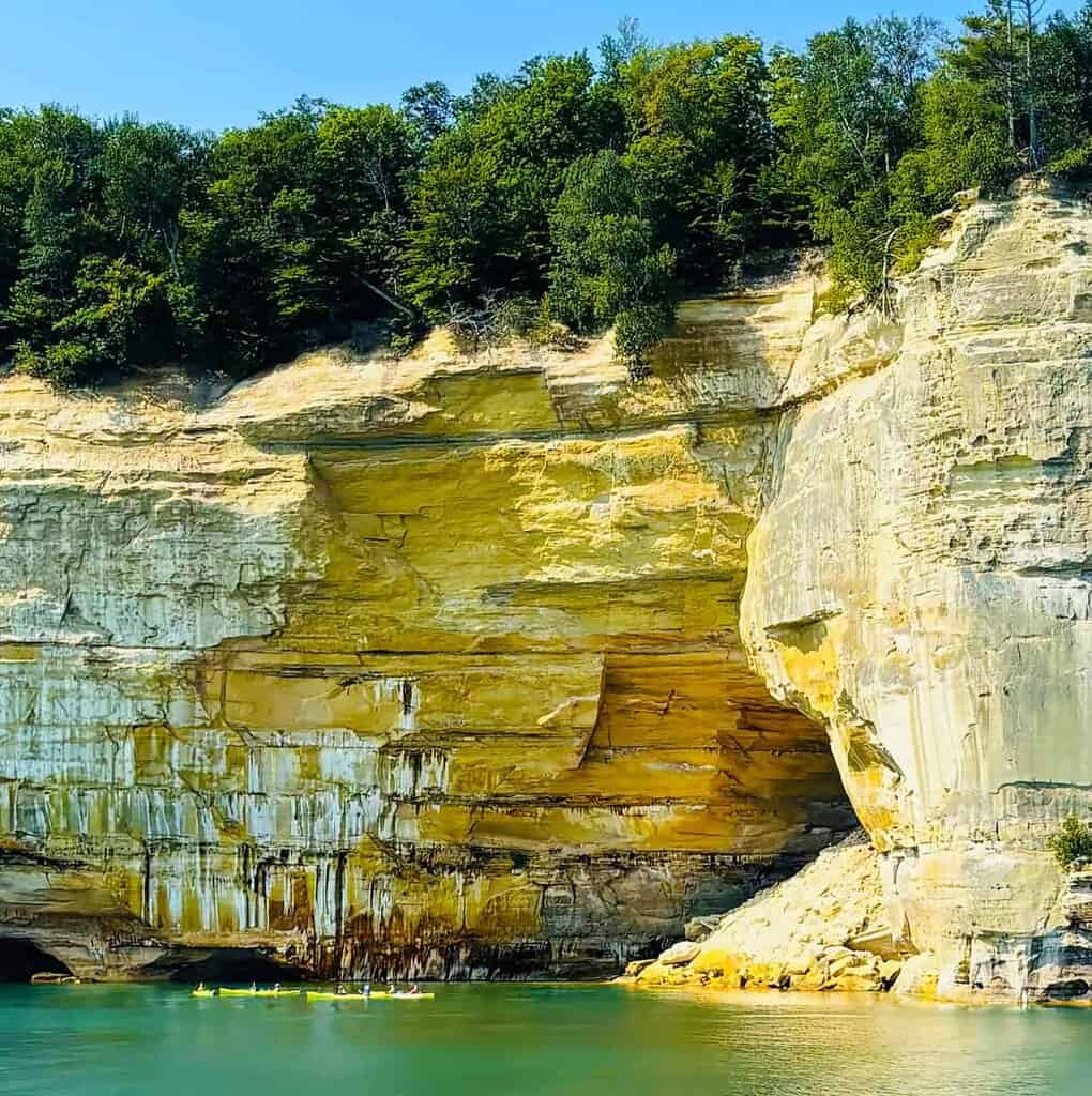 Tall sandstone cliffs with layered colors, green trees on top, and kayakers in the turquoise water below at Pictured Rocks National Lakeshore.