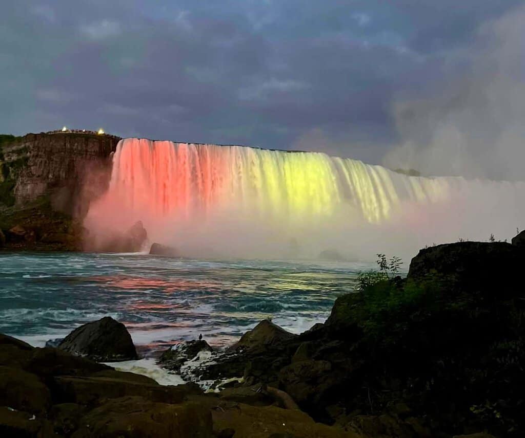 A vibrant waterfall, illuminated in pink, yellow, and orange, cascades down into a misty river against an overcast sky.
