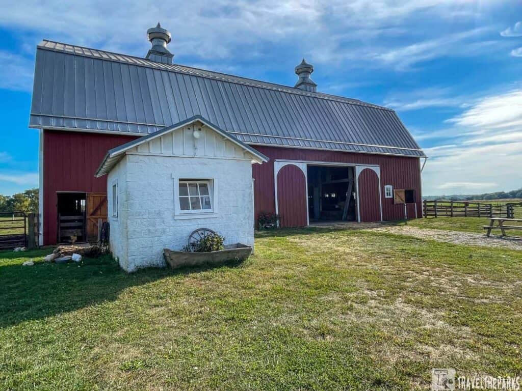 A large red barn with a silver roof next to a small white outbuilding, set in a rural landscape with grass and a blue sky.