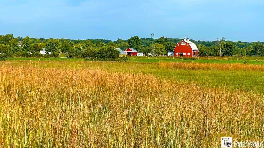 A red barn at the Farm at Prophetstown State Park amidst fields and tall grass with a windmill nearby and trees in the background.

