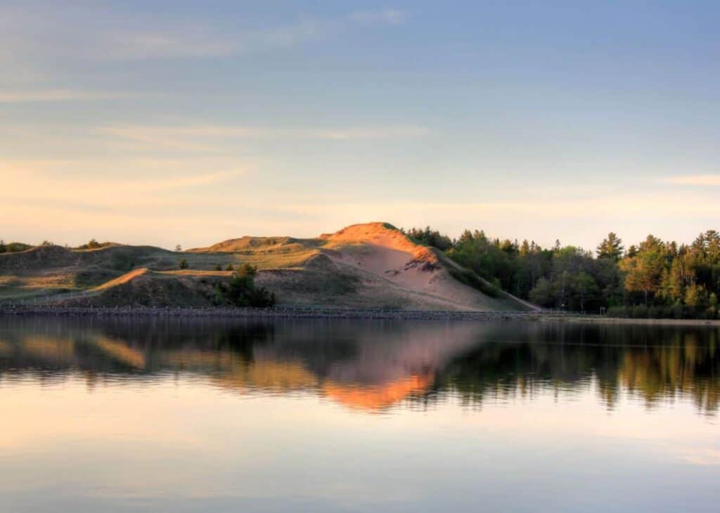 s a serene landscape at Pictured Rocks National Lakeshore, showcasing a sand dune with a pinkish hue during sunset. The dune slopes gently, bordered by dense green foliage on one side. The foreground features a calm body of water reflecting the vibrant colors of the dune and the surrounding greenery. The sky above is clear with subtle gradients of blue and soft orange, indicating a tranquil evening scene.