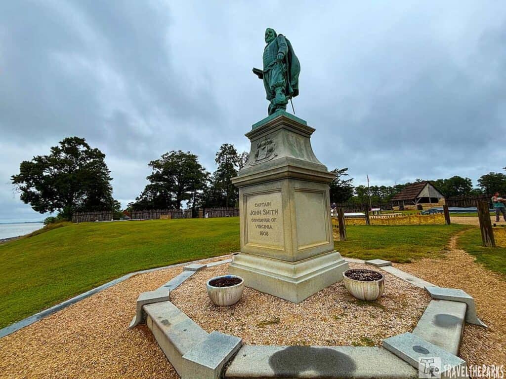 Statue of Captain John Smith on a pedestal in Historic Jamestowne with trees and a historical building in the background.
