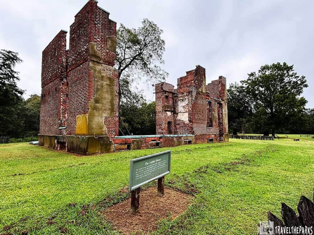 The ruins of Ambler House at Historic Jamestowne, featuring partial red brick walls and a green informational sign, surrounded by grass and trees.


