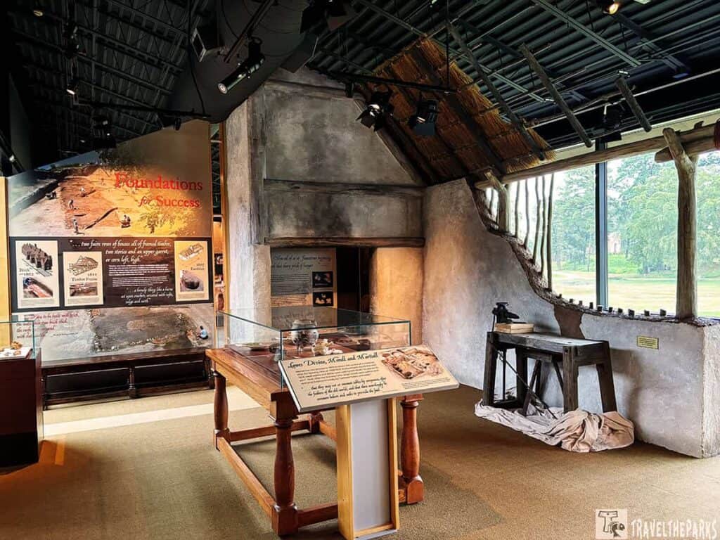 Museum exhibit with informational panels and artifacts in glass cases, adjacent to a thatched roof structure.

