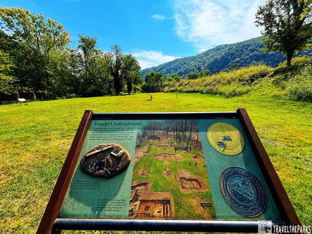 A grassy field at Harpers Ferry armory site with an informational sign in the foreground and tree-covered hills in the background.