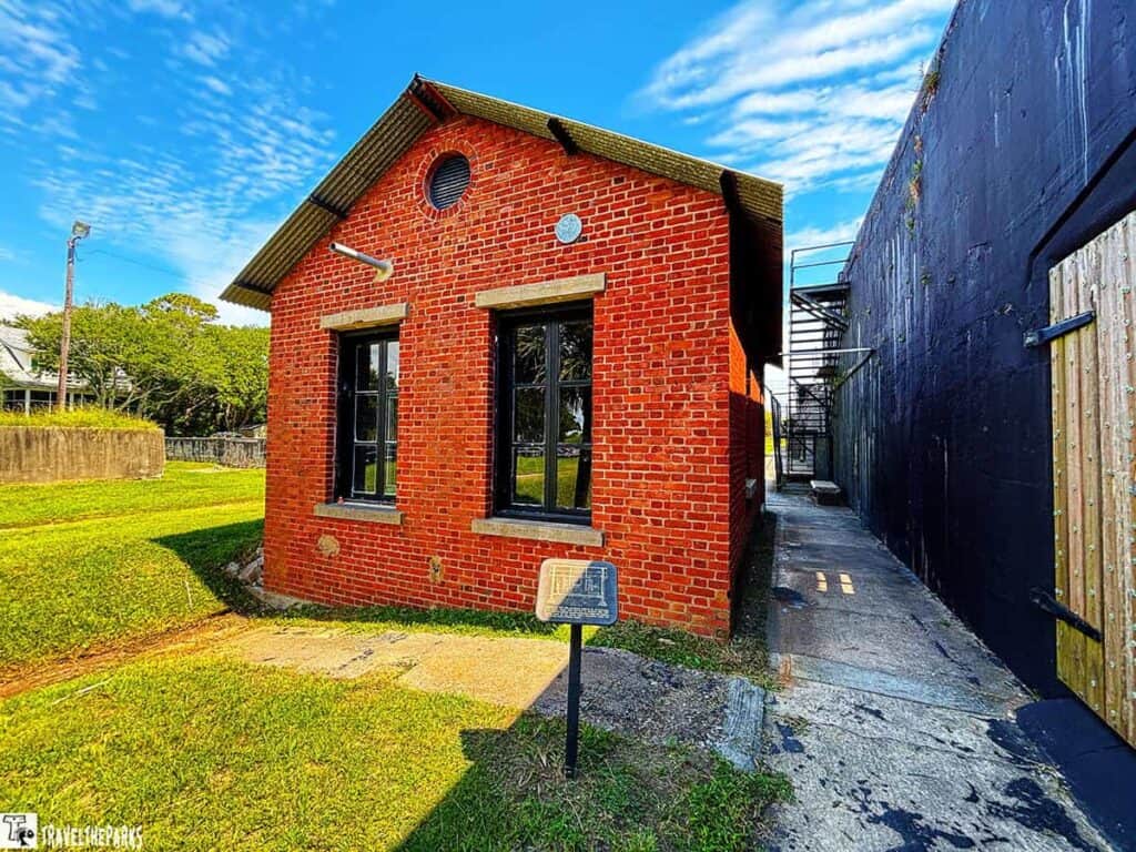 A red brick powerhouse building with black windows and a metal roof, next to a narrow passage and grassy area at Battery Jasper in Fort MOultrie National Monument.

