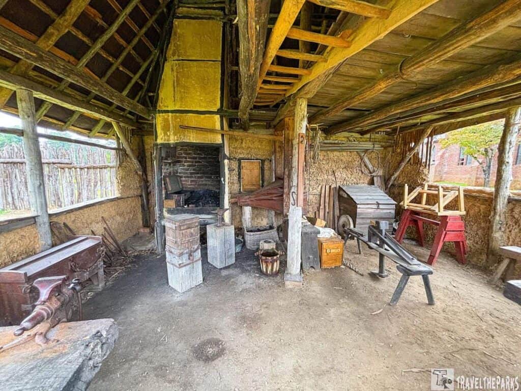 Interior of a historic blacksmith's workshop with a brick hearth, wooden beams, tools, and sunlight filtering through open windows.
