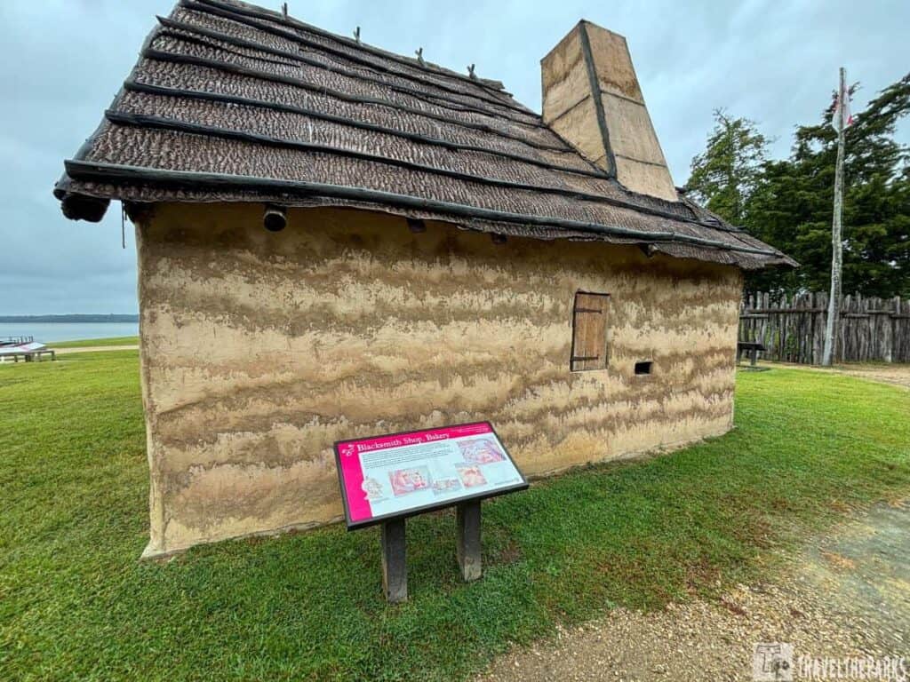 Historic building at Jamestowne with a thatched roof and chimney, labeled as a blacksmith shop and bakery with an informational sign in front.
