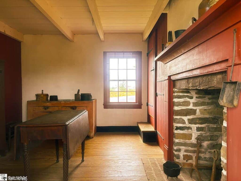 Interior of a rustic room with a stone fireplace, wooden table, cupboard, and a window.