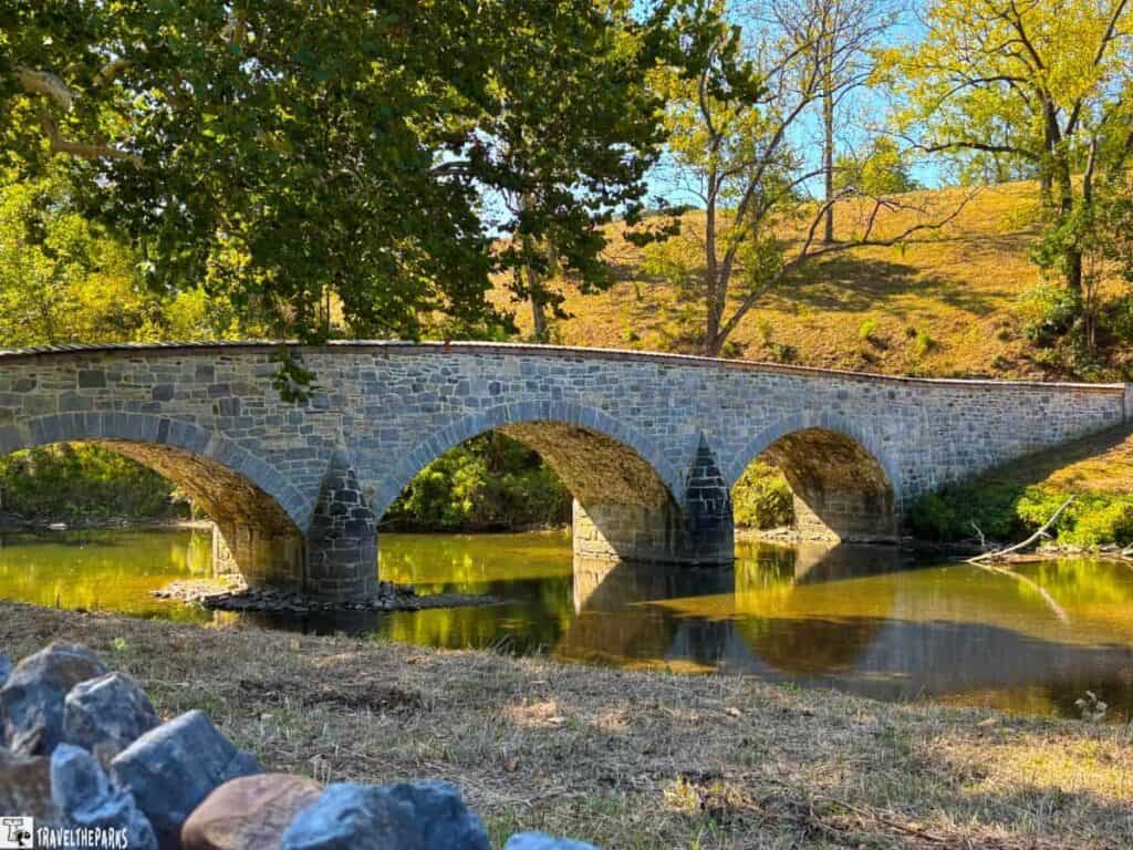 Stone arch Burnside bridge with three arches over a calm river, surrounded by autumnal trees and grassy banks.

