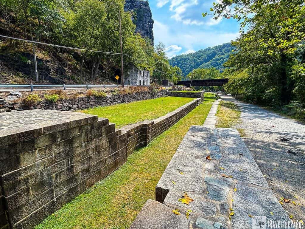 Scenic view of the C&O Canal at Maryland Heights with a stone path, gravel trail, trees, a stone wall, and a hillside.