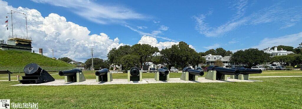 Row of black cannons on display at Fort Moultrie with a grassy mound and flags in the background under a partly cloudy sky.