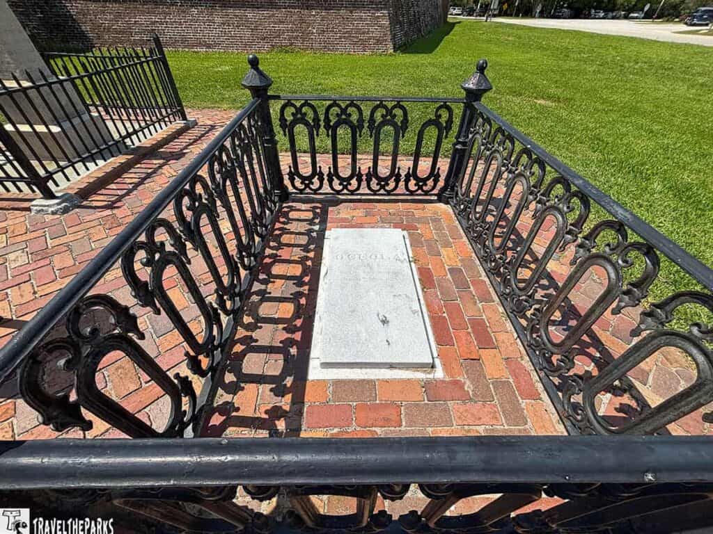 Grave of Chief Osceola enclosed by a decorative black iron fence on a brick path, with grass and brick wall in the background.


