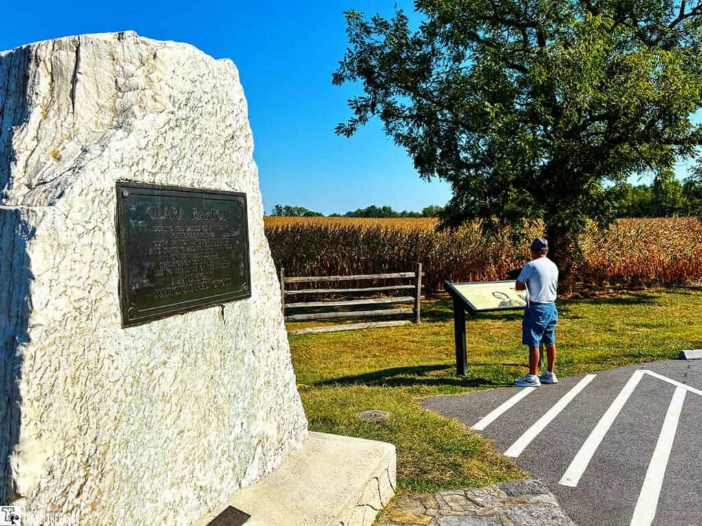 White stone monument for Clara Barton with a bronze plaque; person reading a sign near a cornfield and tree.
