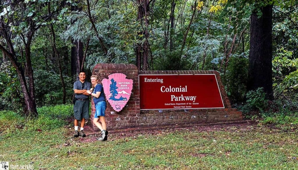 Two people standing by a brick sign for Colonial Parkway, surrounded by dense trees.
